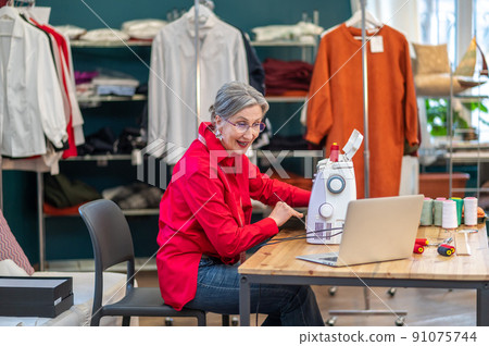 Woman at sewing machine talking at laptop screen Woman at sewing machine talking at laptop screen 91075744