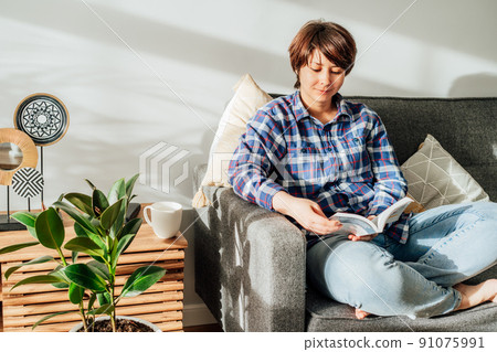Woman sit on the sofa couch and relax while reading a book at home in cozy modern interior living room with green plants. Offline activity lifestyle. Reading hobby. Leisure indoors entertainment 91075991