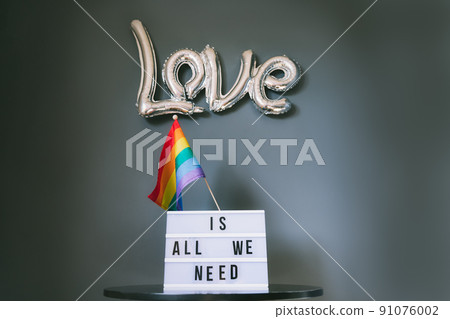 LGBTQ pride month background. A light box with the text Love is all we need under a silver foil balloon with a rainbow flag against a gray wall. Film effect toning. Selective focus. 91076002