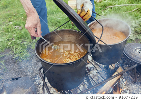 Hiking pot, Bowler in the bonfire. in cauldron at the stake. Traveling, tourism, picnic cooking, cooking at the stake in a cauldron, fire and smoke. Hiking pot, Bowler in the bonfire. in cauldron at the stake. Traveling, tourism, picnic cooking, cooking at the stake in a cauldron, fire and smoke. 91077219