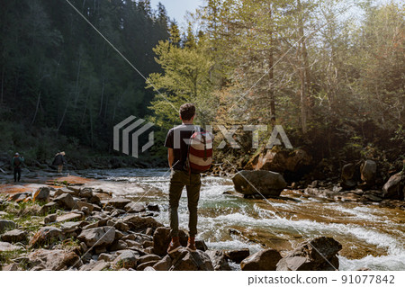 Back view on man hiker with backpack standing at river with rocks and enjoying nature. Back view on man hiker with backpack standing at river with rocks and enjoying nature. 91077842