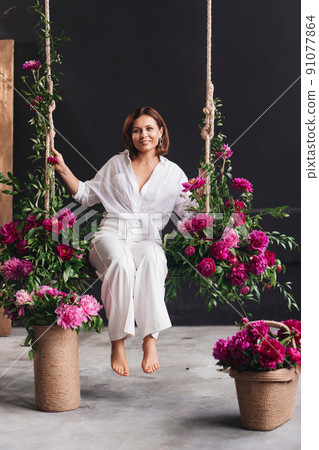 attractive and happy woman in white cozy clothes on a swing with peony flowers 91077864