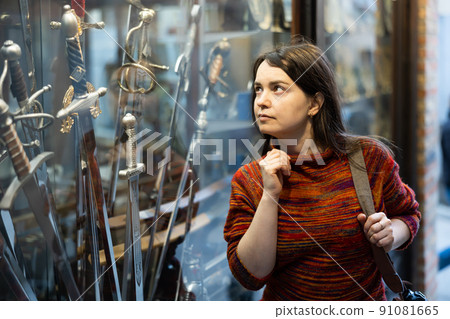 Woman choosing old-fashioned sword in souvenir shop Woman choosing old-fashioned sword in souvenir shop 91081665