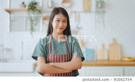 Happy young asian woman wearing apron and standing in kitchen room Happy young asian woman wearing apron and standing in kitchen room 91082354