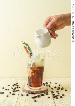 pouring milk in black coffee glass with ice cube, cinnamon and rosemary pouring milk in black coffee glass with ice cube, cinnamon and rosemary 91086147