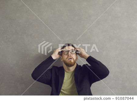 Shocked young man afraid of something looking up sitting against grey background 91087044