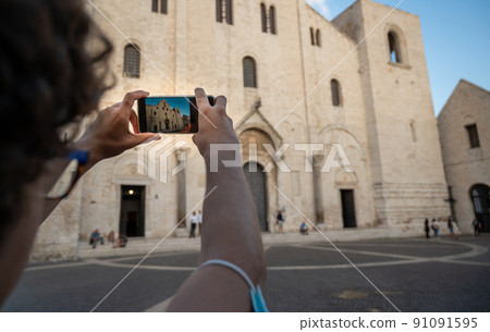 Bari, Puglia, Italy. August 2021. The imposing facade of the church of St. Nicholas, in Roman Catholic style. People in the square. A young man takes a picture with his cellphone 91091595