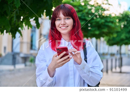 Outdoor portrait of teenage female with smartphone in hands looking at camera, on city Outdoor portrait of teenage female with smartphone in hands looking at camera, on city 91101499