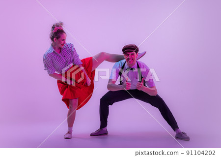 Studio shot of young man and woman in vintage retro style outfits dancing lindy hop isolated on lilac color background in neon light. Studio shot of young man and woman in vintage retro style outfits dancing lindy hop isolated on lilac color background in neon light. 91104202