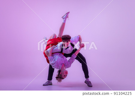 Emotional expressive young man and pretty woman in vintage style outfits dancing retro dance isolated on very peri color background in neon light. Emotional expressive young man and pretty woman in vintage style outfits dancing retro dance isolated on very peri color background in neon light. 91104225