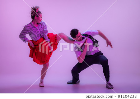 Studio shot of young man and woman in vintage retro style outfits dancing lindy hop isolated on lilac color background in neon light. Studio shot of young man and woman in vintage retro style outfits dancing lindy hop isolated on lilac color background in neon light. 91104238