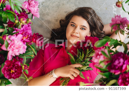 an attractive young woman in a pink blouse with peony flowers on the floor.  91107399