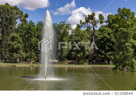 Font. Water fountain in a pond in a Madrid park, with tall green trees in the background on a clear day in Spain. Europe. Horizontal photography. Font. Water fountain in a pond in a Madrid park, with tall green trees in the background on a clear day in Spain. Europe. Horizontal photography. 91108644