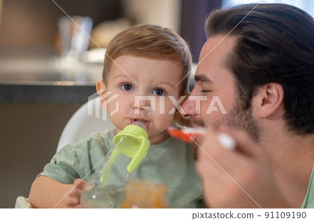 Cute little boy drinking from his bottle while dad feeding him 91109190