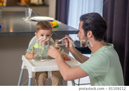Young father feeding his cute baby son and looking funny 91109212