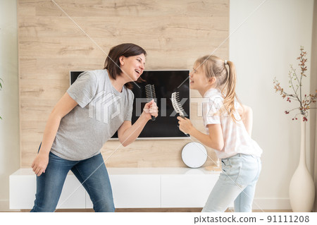 Happy family young adult mother and cute teen daughter having fun singing karaoke song in hairbrushes. mother laughing enjoying funny lifestyle activity with teenage girl at home together. 91111208