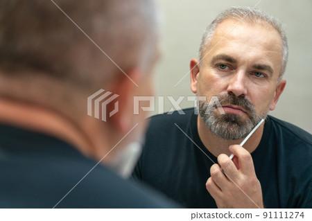 Middle-aged handsome man brushing his beard in the morning in bathroom looking in mirror Middle-aged handsome man brushing his beard in the morning in bathroom looking in mirror 91111274