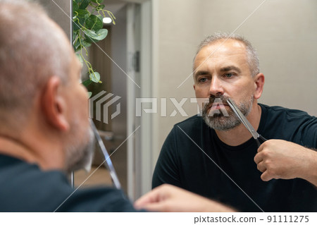 Portrait of Middle-aged handsome man cutting his beard with scissors. Selfcare during quarantine isolation Portrait of Middle-aged handsome man cutting his beard with scissors. Selfcare during quarantine isolation 91111275