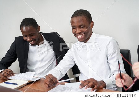 Laughing African American businessmen discussing paperwork with a coworker at a table in an office Laughing African American businessmen discussing paperwork with a coworker at a table in an office 91111417