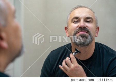 Charming middle aged man shaving his beard and moustache off in front of a mirror to look younger Charming middle aged man shaving his beard and moustache off in front of a mirror to look younger 91111620