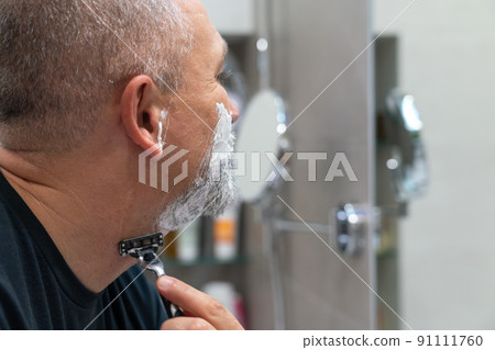 Middle aged handsom man trimming his beard and mustache standing in bathroom and looking in a mirror Middle aged handsom man trimming his beard and mustache standing in bathroom and looking in a mirror 91111760