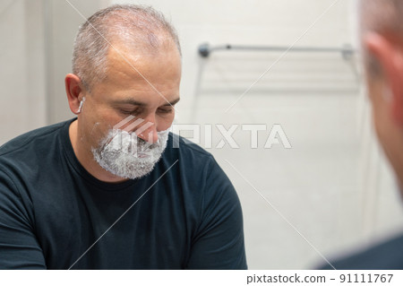 Handsome white-haired beared man shaving off his beard looking in a mirror. Concept of changing style Handsome white-haired beared man shaving off his beard looking in a mirror. Concept of changing style 91111767
