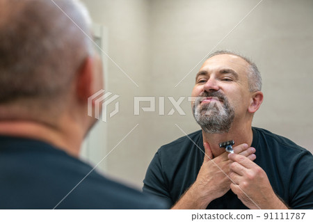 Serious handsome man looking in mirror and shaving his whiskers 91111787