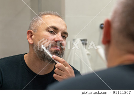 Middle-aged handsome man brushing his beard in the morning in bathroom looking in mirror 91111867