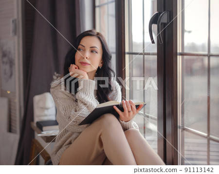 Beautiful young woman reading book near window at home 91113142