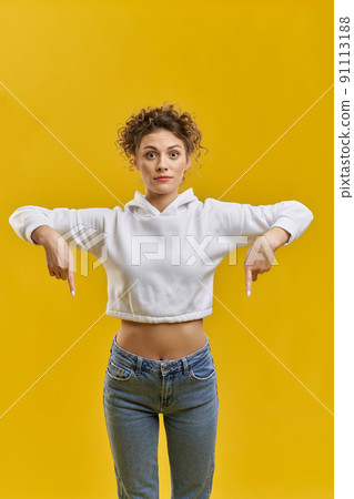 Beautiful stylish woman pointing with fingers down, standing inside. Front view of slim girl showing down direction with fingers gesture, isolated on orange studio background. Concept of gesturing. Beautiful stylish woman pointing with fingers down, standing inside. Front view of slim girl showing down direction with fingers gesture, isolated on orange studio background. Concept of gesturing. 91113188