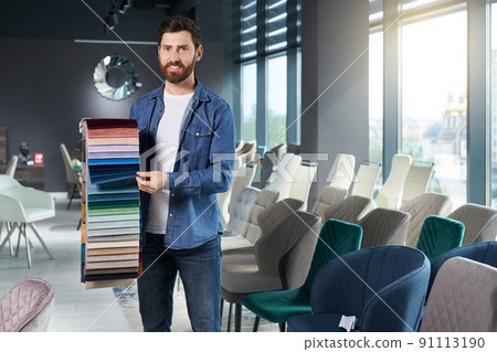 Professional male decorator presenting various samples of upholstery fabric in furniture store. Front view of strong man showing colorful palette, while smiling at camera. Concept of interior design. Professional male decorator presenting various samples of upholstery fabric in furniture store. Front view of strong man showing colorful palette, while smiling at camera. Concept of interior design. 91113190