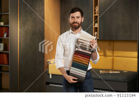 Confident male expert presenting variety samples of upholstery fabric in dark lightened showroom. Portrait of smiling brunet salesman showing colorful textile palette. Concept of interior design. Confident male expert presenting variety samples of upholstery fabric in dark lightened showroom. Portrait of smiling brunet salesman showing colorful textile palette. Concept of interior design. 91113194