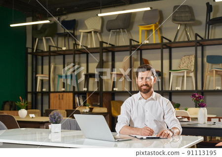 Smiling manager sitting at table with laptop, while working remotely. Front view of confident retailer looking at camera, while writing against rack with chairs on background. Concept of workspace. Smiling manager sitting at table with laptop, while working remotely. Front view of confident retailer looking at camera, while writing against rack with chairs on background. Concept of workspace. 91113195