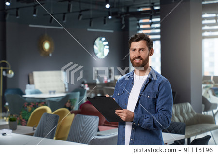 Successful store owner in denim shirt making notes, while working in modern furniture shop. Portrait of handsome designer with tablet smiling at camera, while standing in expo center. Concept of work. Successful store owner in denim shirt making notes, while working in modern furniture shop. Portrait of handsome designer with tablet smiling at camera, while standing in expo center. Concept of work. 91113196