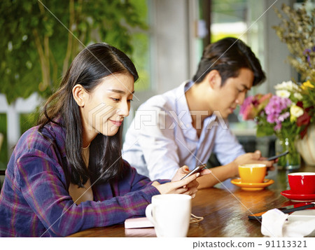 two young asian adults man and woman sitting in coffee shop playing with mobile phone two young asian adults man and woman sitting in coffee shop playing with mobile phone 91113321