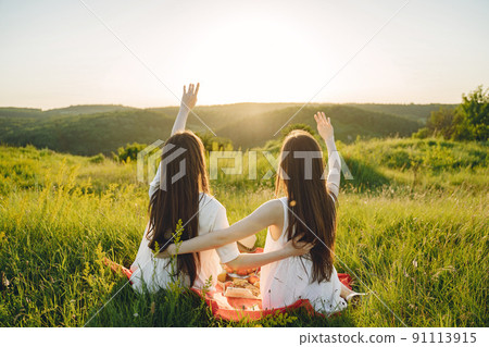 Two pretty sisters in a field of wheat. Beautiful two girls with long healthy hair enjoying nature on summer. Girls on a picnic. 91113915