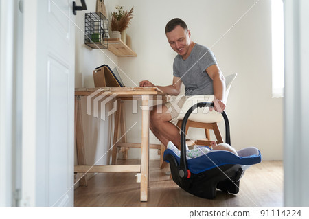A handsome freelance man works on a modern laptop. A young father works at home and cradles his little son in a cradle. High quality photo A handsome freelance man works on a modern laptop. A young father works at home and cradles his little son in a cradle. High quality photo 91114224
