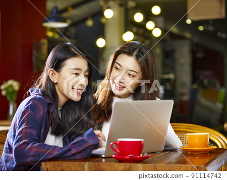 two happy young asian women sitting in coffee shop looking at laptop computer together 91114742