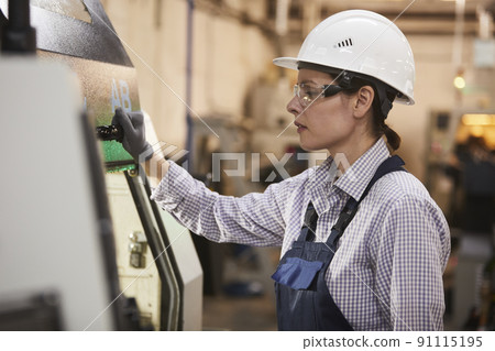 Concentrated gritty young woman in hardhat and safety goggles preparing detail for processing and closing cover of cnc machine at factory 91115195