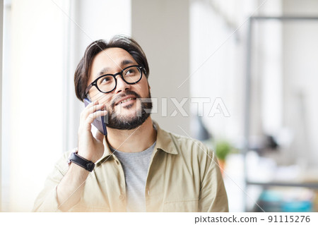 Positive pensive young Asian man with beard sitting by window and using mobile phone for effectively communication 91115276