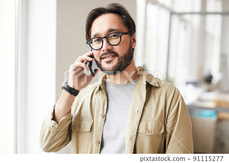 Portrait of satisfied young Asian man with beard calling by cell phone while working in office 91115277