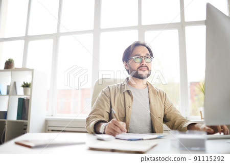 Serious thoughtful young Asian web designer with beard sitting at desk and making note while working with computer in office 91115292