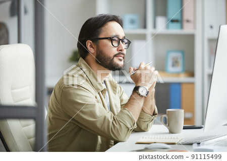 Serious pensive young Asian man with beard and mustache sitting at desk in office and looking at computer monitor while analyzing web design 91115499