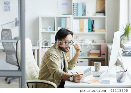Serious busy young Asian man in casual shirt sitting at table with computer and viewing sketches in notepad 91115506