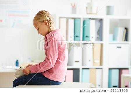 Serious scared little girl with blond braids sitting on examination table in doctors office and nervously strumming toy 91115535