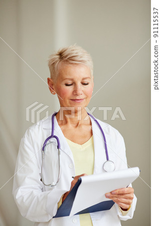 Content beautiful female doctor in lab coat wearing stethoscope around neck filling medical record during examination Content beautiful female doctor in lab coat wearing stethoscope around neck filling medical record during examination 91115537