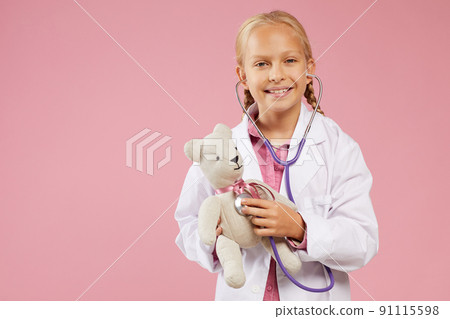 Portrait of happy beautiful little girl with blond braids wearing white coat using stethoscope while examining toy bear 91115598