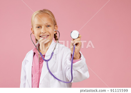 Portrait of happy little girl in lab coat standing against pink background and using stethoscope while dreaming to become doctor 91115599