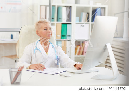 Content confident attractive lady doctor with short hair sitting at table and using computer while working with papers Content confident attractive lady doctor with short hair sitting at table and using computer while working with papers 91115792