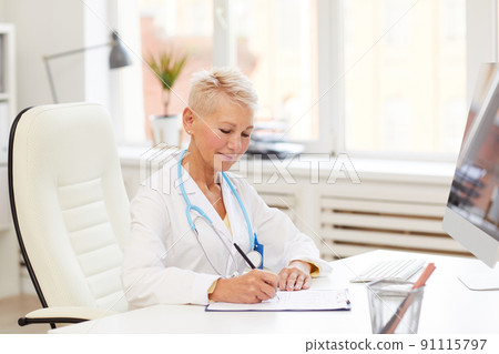 Smiling beautiful mature doctor with stethoscope sitting at desk and adding information about patient in medical record while working in own office Smiling beautiful mature doctor with stethoscope sitting at desk and adding information about patient in medical record while working in own office 91115797